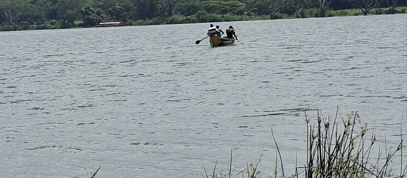 Three people in a wooden canoe on the calm open water of Lake Victoria, green shoreline in the background